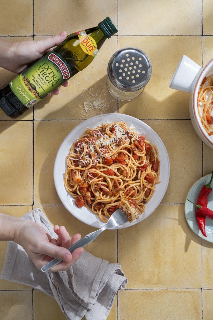 Tanier špagiet alla Amatriciana posypaný strúhaným syrom na žltom keramickom povrchu, vedľa fľaše Borges extra panenského olivového oleja, so soľničkou a červenou čili papričkou na tanieriku. Ruka držiaca vidličku sa chystá nabrať kúsok cestovín.